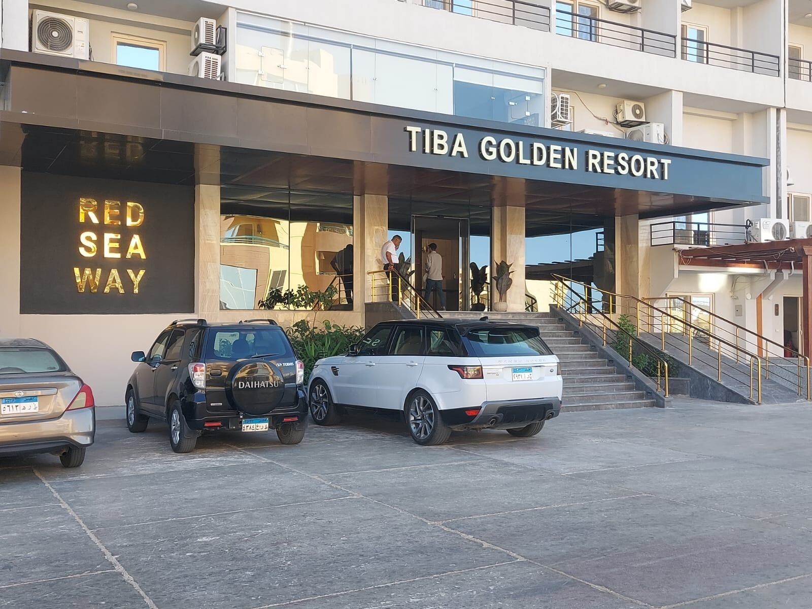 A building with a sign reading 'TIBA GOLDEN RESORT' and a 'RED SEA WAY' sign. There are two cars parked in front.
