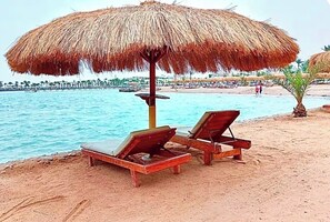A beach with two wooden lounge chairs under a thatched umbrella, a sandy shore, and a calm sea. - Serenity Sands Red Sea Hurghada (Red Sea Governorate)