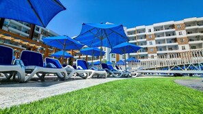 A row of lounge chairs under blue umbrellas, a grassy area, and a building with balconies. - Serenity Sands Red Sea Hurghada (Red Sea Governorate)