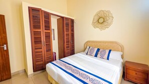 A bedroom with a bed, a wooden wardrobe, a nightstand, and a decorative wall piece. - Adia Gardens, Shanzu, Mombasa (Mombasa)
