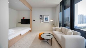 A modern hotel room with a bed, a sofa, a small table, and a large window offering a city view. - Modern Upside Studio w Skyline View (Dubai)