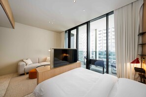 Ein modernes Hotelzimmer mit einem großen Bett, einer Couch, einem Fernseher und Blick auf die Stadt durch raumhohe Fenster.