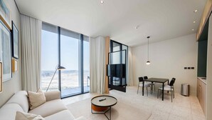 A modern living room with a sofa, a coffee table, a dining area with a table and chairs, a TV, and large windows offering a view. - Prime Upside Studio w Private Balcony (Dubai)