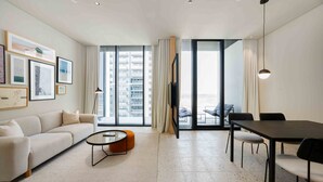 A modern living room with a sofa, coffee table, dining table, and chairs. There are large windows with curtains, a flat-screen TV, and artwork on the wall. - Prime Upside Studio w Private Balcony (Dubai)