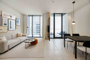 A modern living room with a sofa, coffee table, dining table, and chairs. There are large windows with curtains, a flat-screen TV, and artwork on the wall. - Prime Upside Studio w Private Balcony (Dubai)