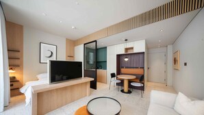 Ett modernt hotellrum med en platt-TV, en soffa, ett runt bord och en liten orange pall.