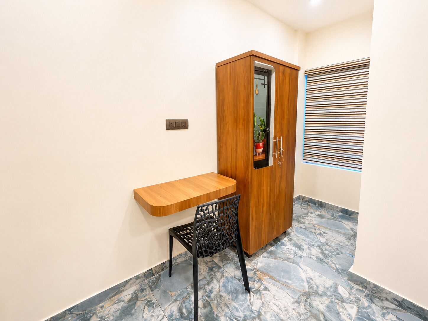 A small room with a wooden desk, a chair, a wardrobe, and a window with blinds.