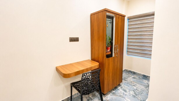 Ein kleines Zimmer mit einem Holztisch, einem Stuhl, einem Kleiderschrank und einem Fenster mit Jalousien.