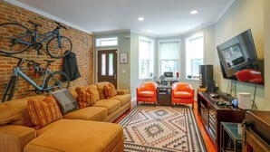 A living room with a brick wall, a sofa, a coffee table, two armchairs, a television, and a bicycle. - Quiet Home in Walkable Area: 3 Mi to National Mall (Washington)