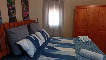 A bedroom with a bed, pillows, a blanket, a wooden wardrobe, and a window with curtains.