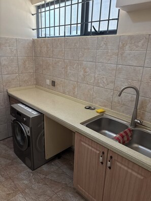 Une cuisine compacte équipée d’un lave-linge encastré, d’un évier avec un robinet et d’une fenêtre avec des barreaux.