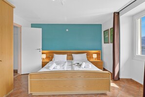Ein Schlafzimmer mit einem hölzernen Bett, weißer Bettwäsche, einer Türkis akzentuierten Wand, einem Fenster mit Ausblick und einer Tür zu einem anderen Raum.