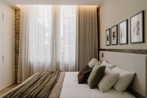 A bedroom with a bed, a headboard, a wall-mounted light, and a window with curtains. - Fulham Residences Two (London)