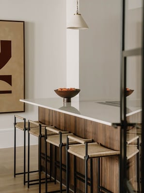 A modern kitchen with a white countertop, wooden bar stools, and a hanging pendant light. - Fulham Residences Two (London)