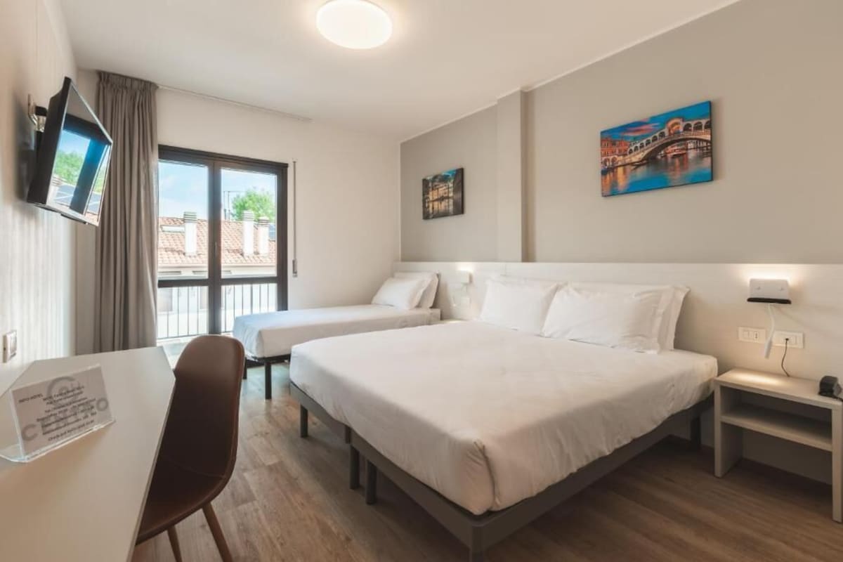 Hotellihuone, jossa on kaksi sänkyä, työpöytä, tuoli, televisio ja taideteoksia seinillä.