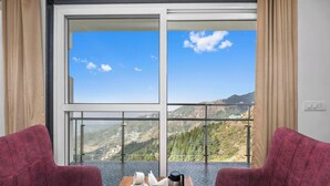 A room with two red chairs facing a large window with a mountain view. A wooden table with a tray of cookies and a coffee pot is in the center. - The Palm Aryan Mussoorie 5min Mall Road (Mussoorie)