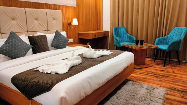 A hotel room with a large bed, two chairs, a small table, and a wooden nightstand. - The Palm Aryan Mussoorie 5min Mall Road (Mussoorie)
