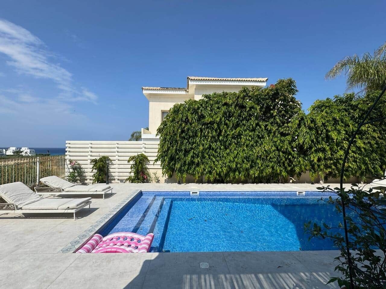 Une piscine avec des chaises longues, un ciel dégagé et un bâtiment à la façade blanche, orné de verdure.