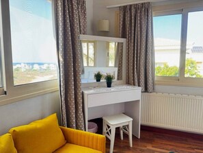 Une pièce avec un bureau blanc, un petit tabouret, un canapé jaune et une vue sur l’extérieur grâce à de grandes fenêtres.