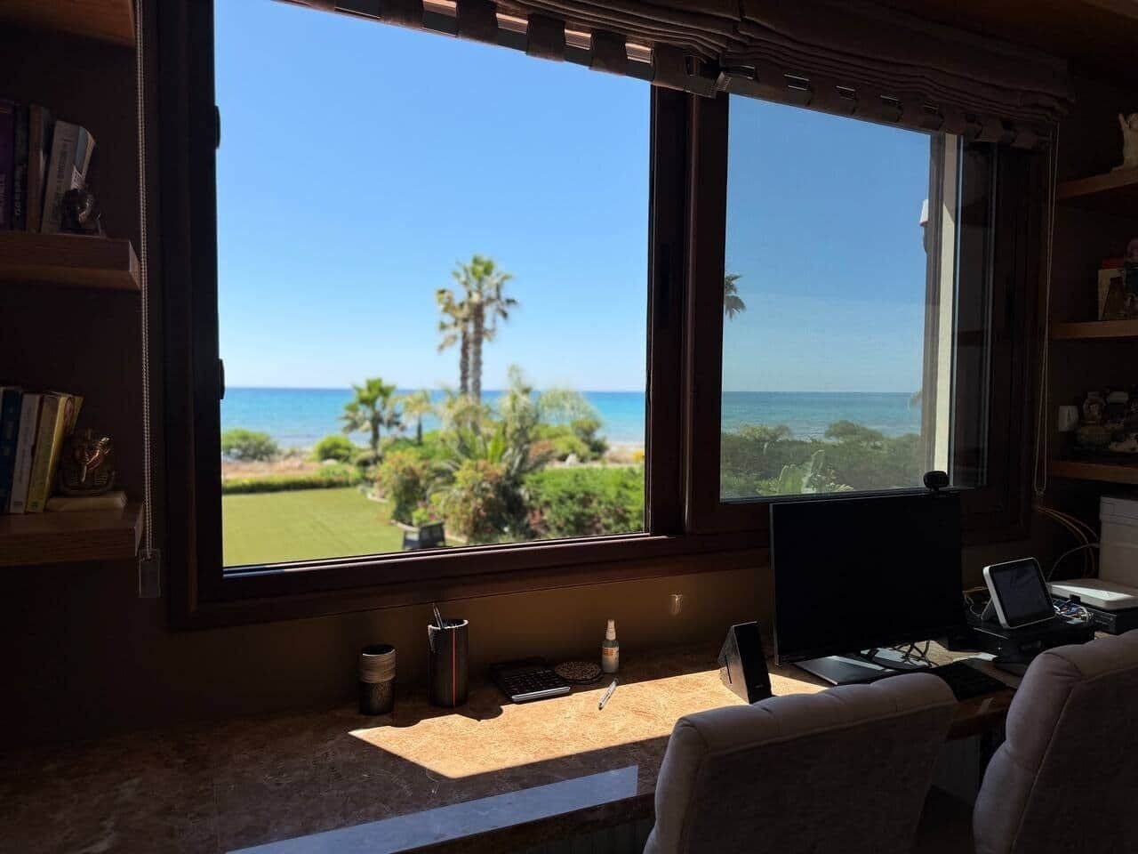 A room with a desk, a computer, and a large window overlooking a beach and ocean.