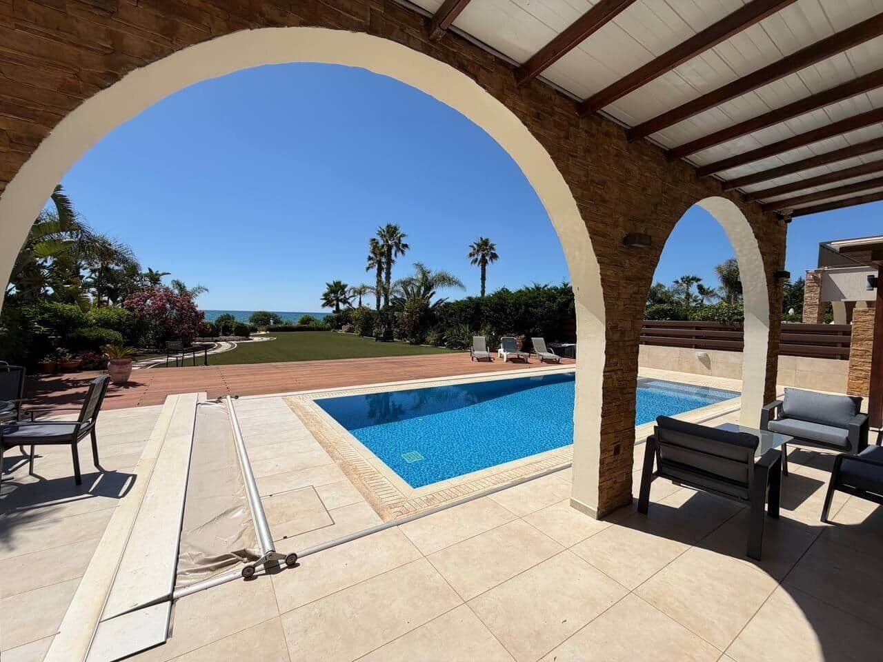 A covered outdoor swimming pool area with a view of the ocean, lounge chairs, and a pergola structure.