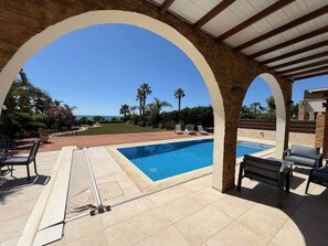 Ein überdachter Poolbereich im Freien mit Blick auf das Meer, Liegestühle und eine Pergola.