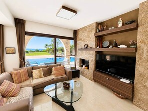 A modern living room with a sofa, a glass coffee table, a flat-screen TV, and a pool view. - Emerald Seafront Residence (Sotira)