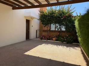 A covered outdoor area with a gravel floor, a wooden door, and a garden with flowering plants and a fence. - Emerald Seafront Residence (Sotira)
