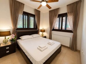 Ein Hotelzimmer mit einem Bett, zwei Nachttischlampen, einem Nachttisch, einem Fenster mit Jalousien und einem Deckenventilator.