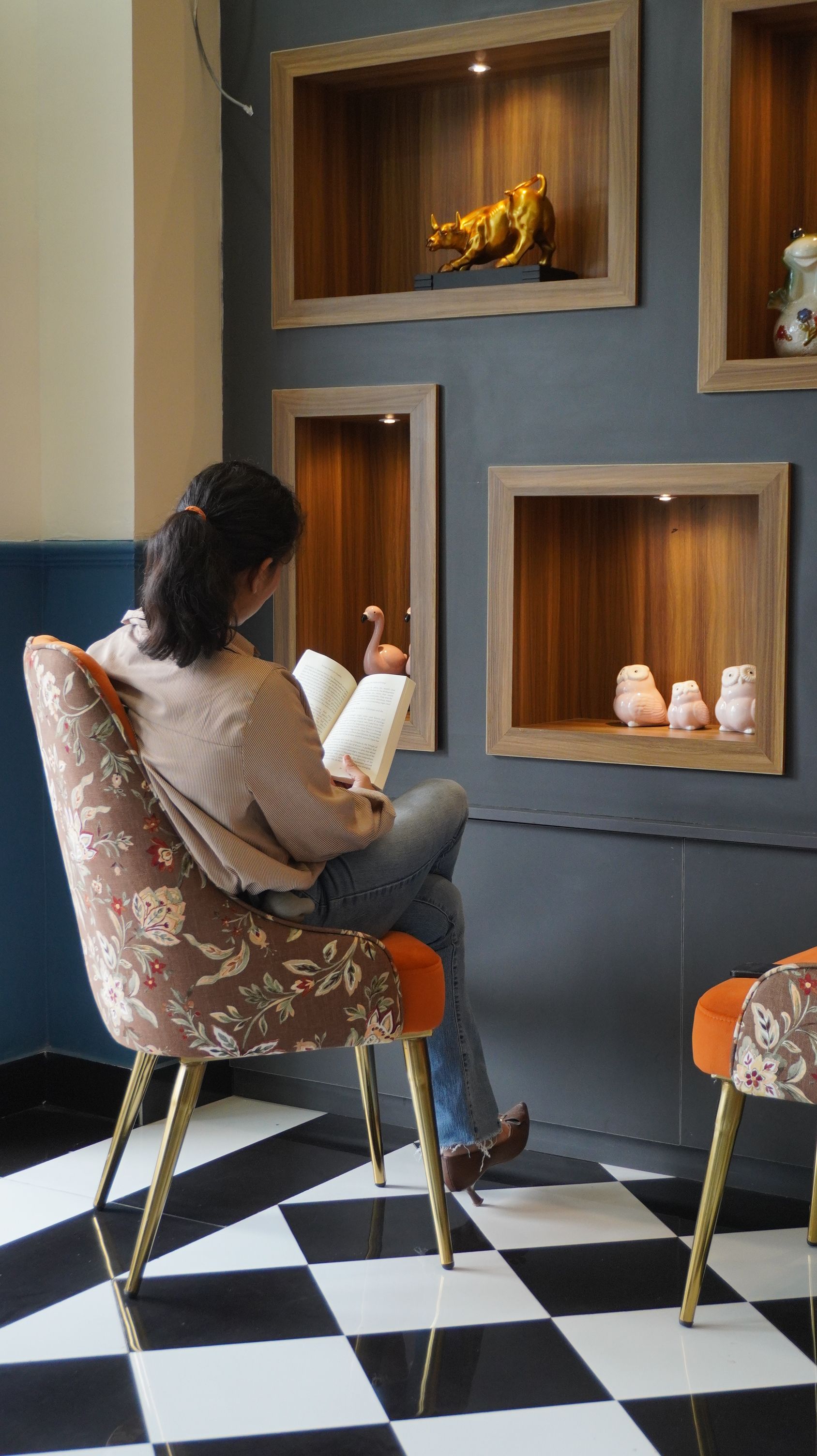 A person seated on a floral-patterned chair reading a book in a room with checkered flooring, wooden shelves with decorative items, and a wall-mounted light fixture.