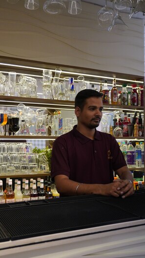 Una persona de pie detrás de una barra con varios vasos y botellas al fondo.