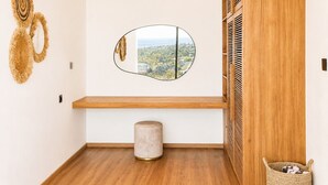 Ein Zimmer mit einer Holzbank, einem runden Spiegel der eine Stadtansicht reflektiert, einem kleinen Hocker und einem gewebten Korb.
