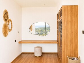Ein Zimmer mit einer Holzbank, einem runden Spiegel der eine Stadtansicht reflektiert, einem kleinen Hocker und einem gewebten Korb.