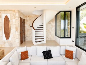 A modern interior with a stone wall, a curved staircase, a white sofa with decorative pillows, and a large window with a view.