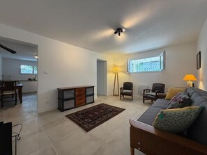 A spacious living room with a sofa, chairs, a coffee table, and a rug. There is a window with a view, a lamp, and a ceiling fan. - Kanbo's Getaway - Downtown (CAMBO LES BAINS)