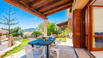 A covered outdoor seating area with a table and chairs, stone flooring, and a view of the surrounding landscape.