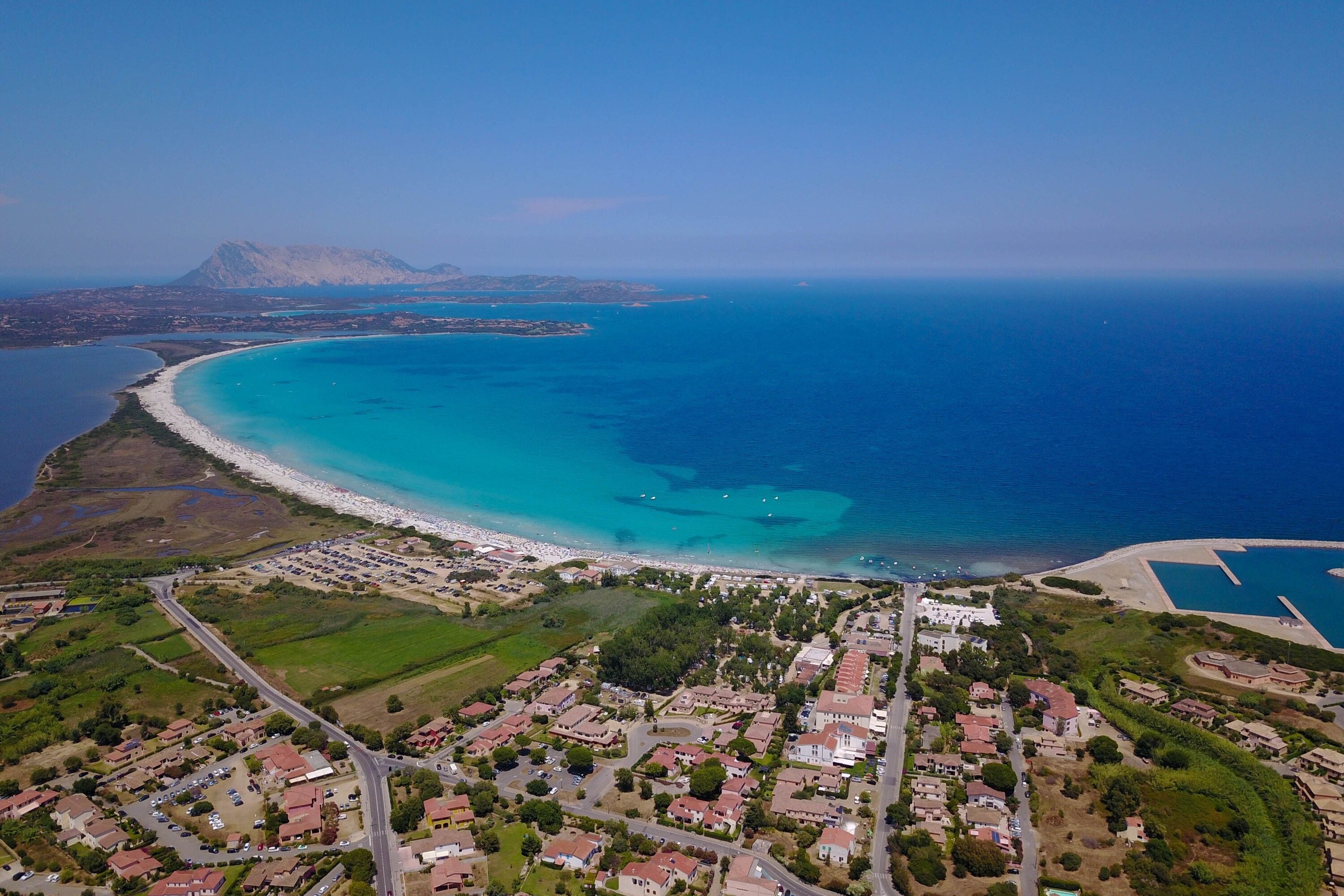 Vue aérienne d’une ville côtière avec une plage, des zones résidentielles et un paysage montagneux en arrière-plan.