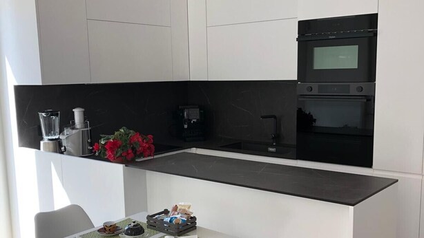 Una cocina moderna con armarios blancos, encimera negra y horno empotrado. Hay una pequeña zona de comedor con una mesa y dos sillas.