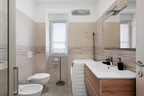 A modern bathroom with a toilet, bidet, vanity with a mirror, and a window.