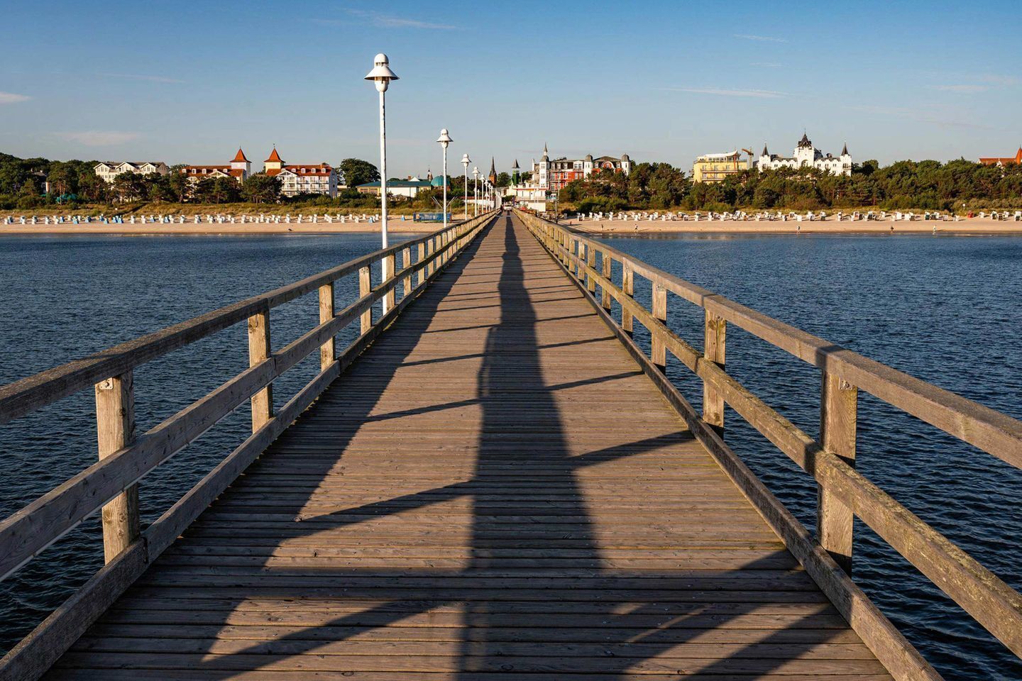 A wooden pier extending into calm water, with buildings and a clear sky in the background.