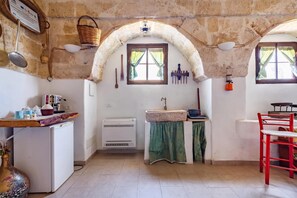 A compact kitchen with a refrigerator, a sink, a window with curtains, a hanging light fixture, and a red chair. - Masseria Le Mesolecchie - Ulivo Apt (Crispiano)