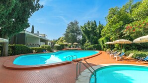 A swimming pool with lounge chairs and umbrellas, surrounded by trees and a brick pathway. - Hendrik Vittoriale Lake View Shared Pool (Gardone Riviera)
