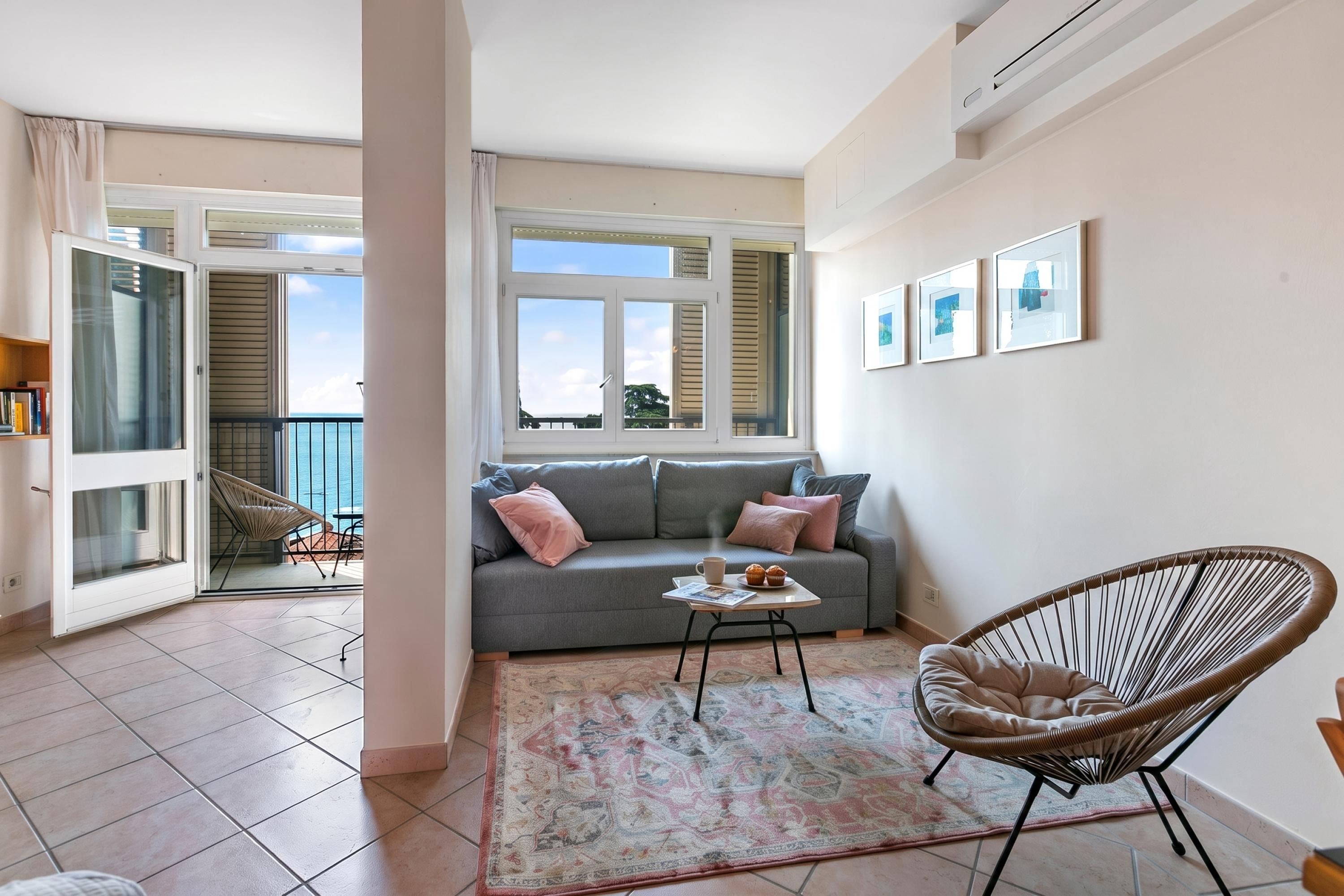 A living room with a grey sofa, a wicker chair, a coffee table, and a view of the sea through large windows.