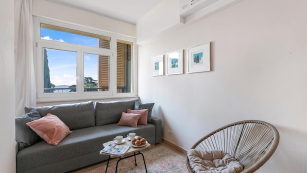 A modern living room with a grey sofa, a wicker chair, and a window with blinds. - Hendrik Vittoriale Lake View Shared Pool (Gardone Riviera)