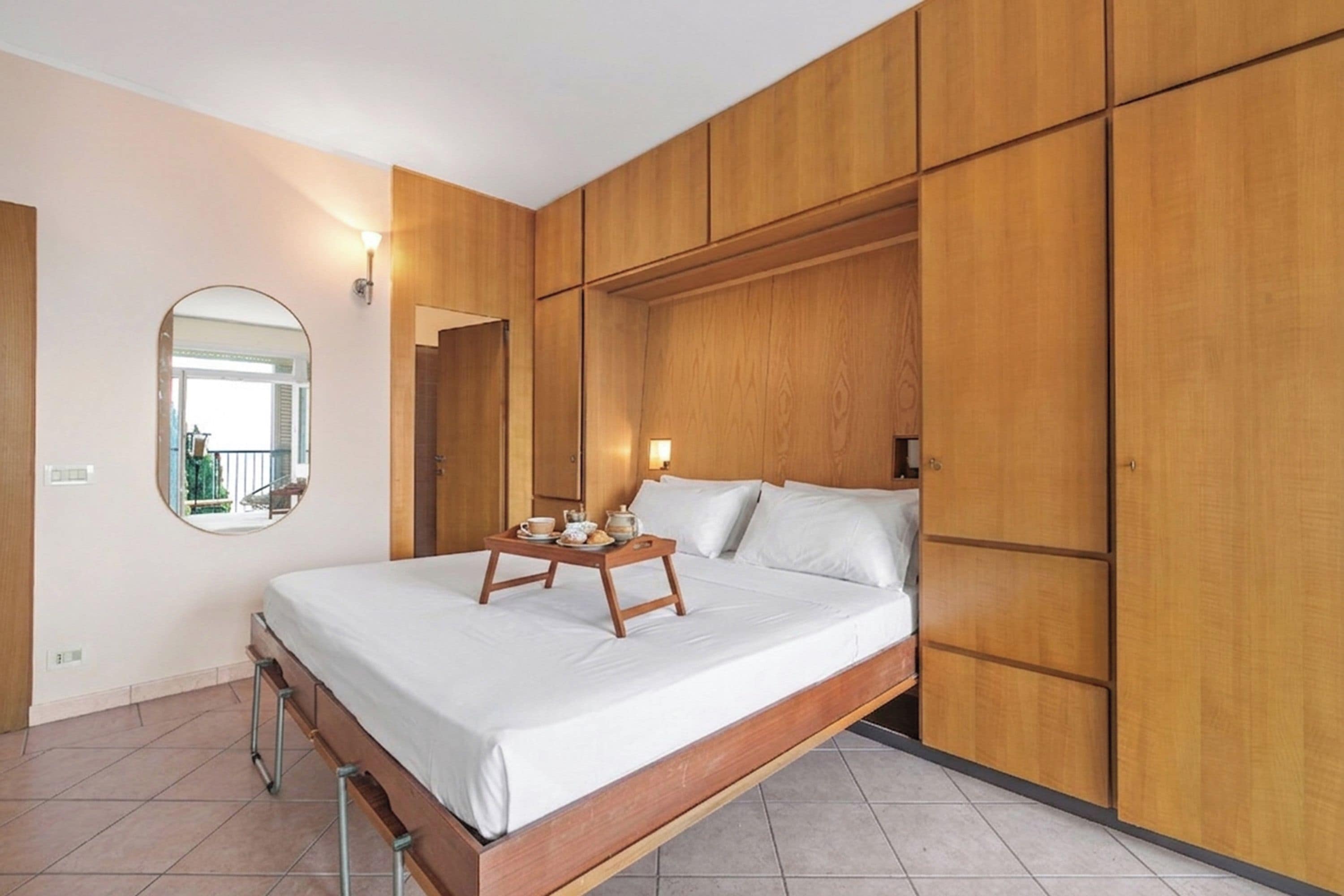 Een hotelkamer met een bed, een houten garderobe, een ronde spiegel en een dienblad met ontbijtartikelen.