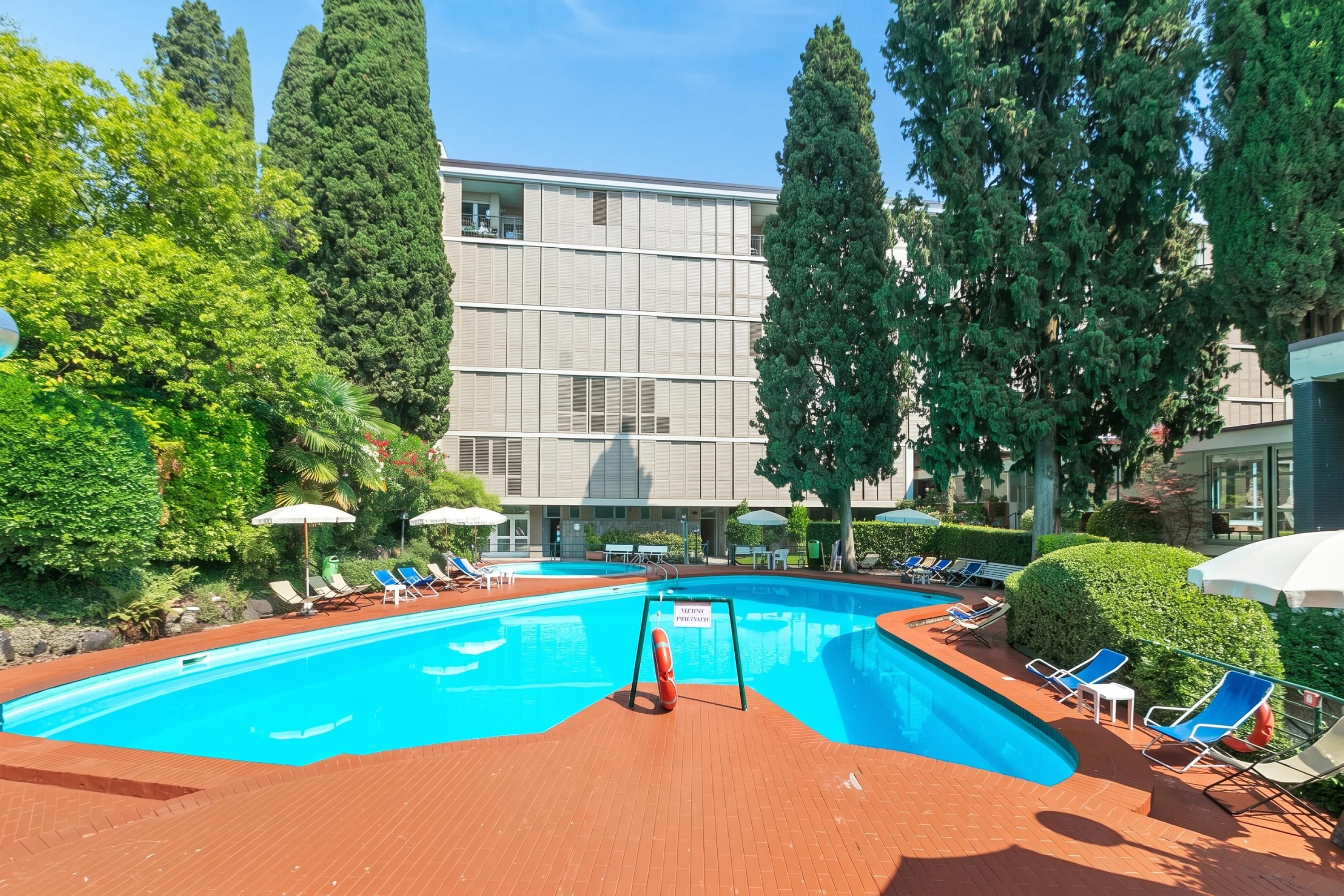 A swimming pool with lounge chairs and umbrellas, surrounded by trees and a building with large windows.