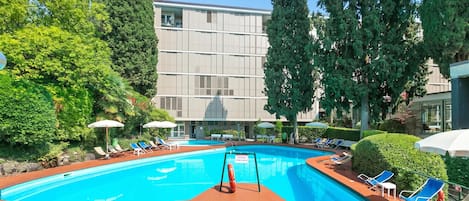 A swimming pool with lounge chairs and umbrellas, surrounded by trees and a building with large windows.