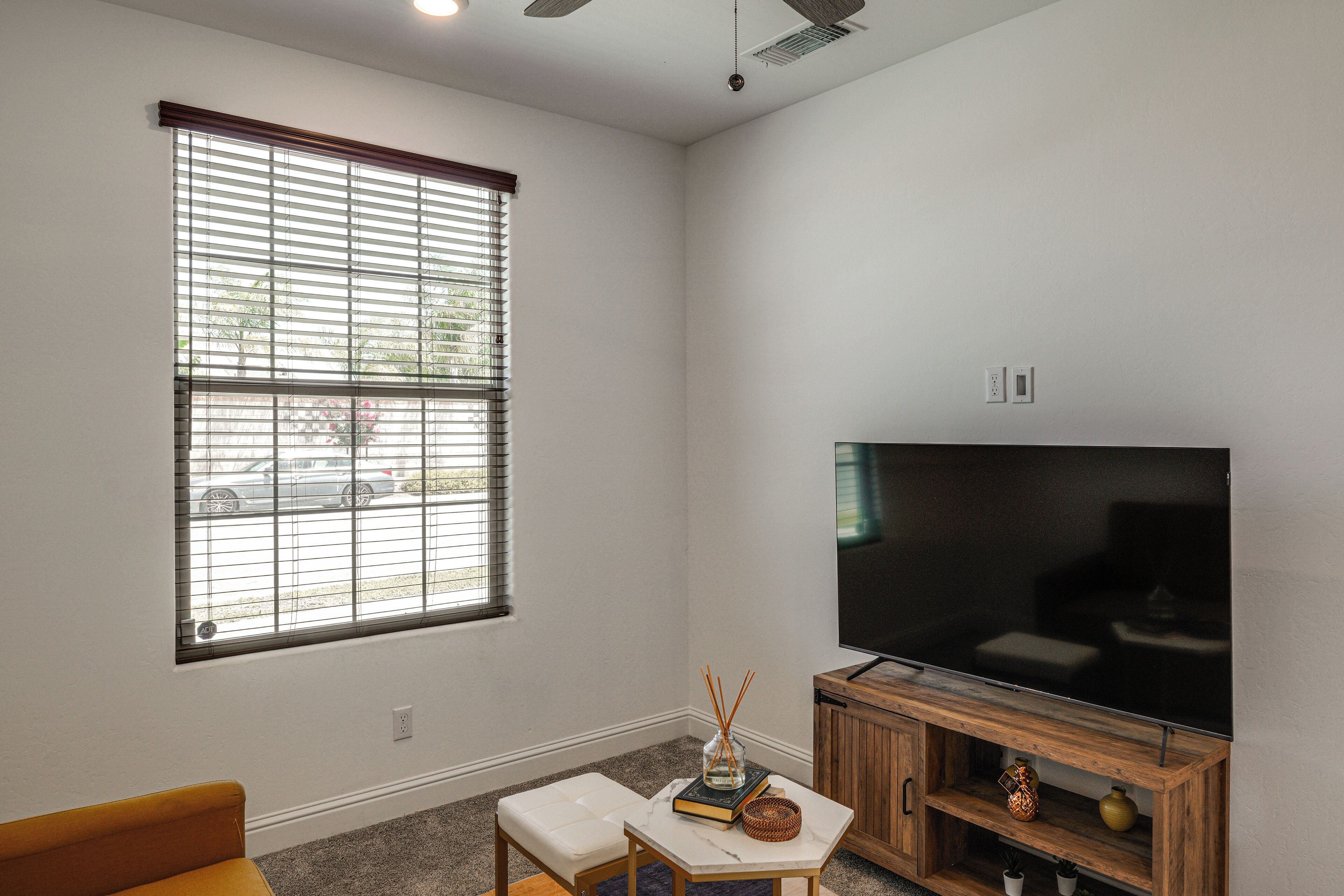 A living room with a large flat-screen TV, a window with blinds, a small table with a vase, and a wooden cabinet.
