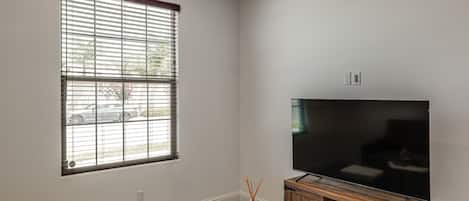 Un salón con un televisor de pantalla plana grande, una ventana con persianas, una mesita con un jarrón y un mueble de madera.