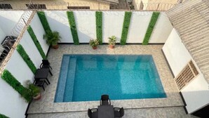 Una piscina rectangular con un borde empinado de baldosas, rodeada de muros blancos con detalles verticales verdes y plantas en macetas.